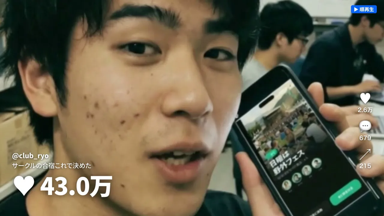 最終版の大学生。蛍光灯の部室で話しかけている途中の表情。後ろで学生が作業している